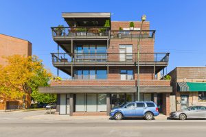 Three-story brick building with balconies and a blue SUV parked in front.