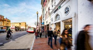 People strolling along a brick sidewalk in front of shops on a city street.
