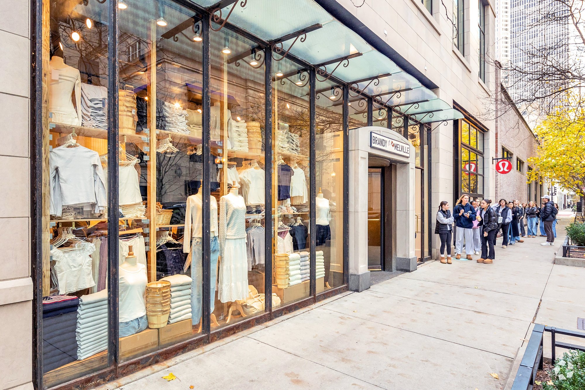 Exterior view of a clothing store storefront with mannequins and customers waiting in line.
