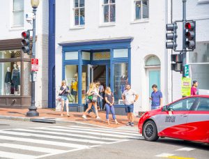 People walking near a storefront and a red car at a crosswalk.
