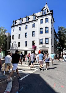 A crowd of pedestrians crossing a street in front of a large white building.