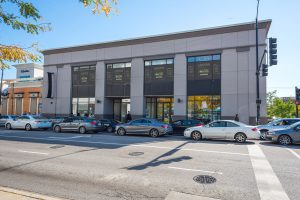 Street-level view of a long, low building with multiple storefront windows and cars parked in front.