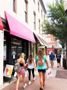 People walking on a city sidewalk past shops with awnings.