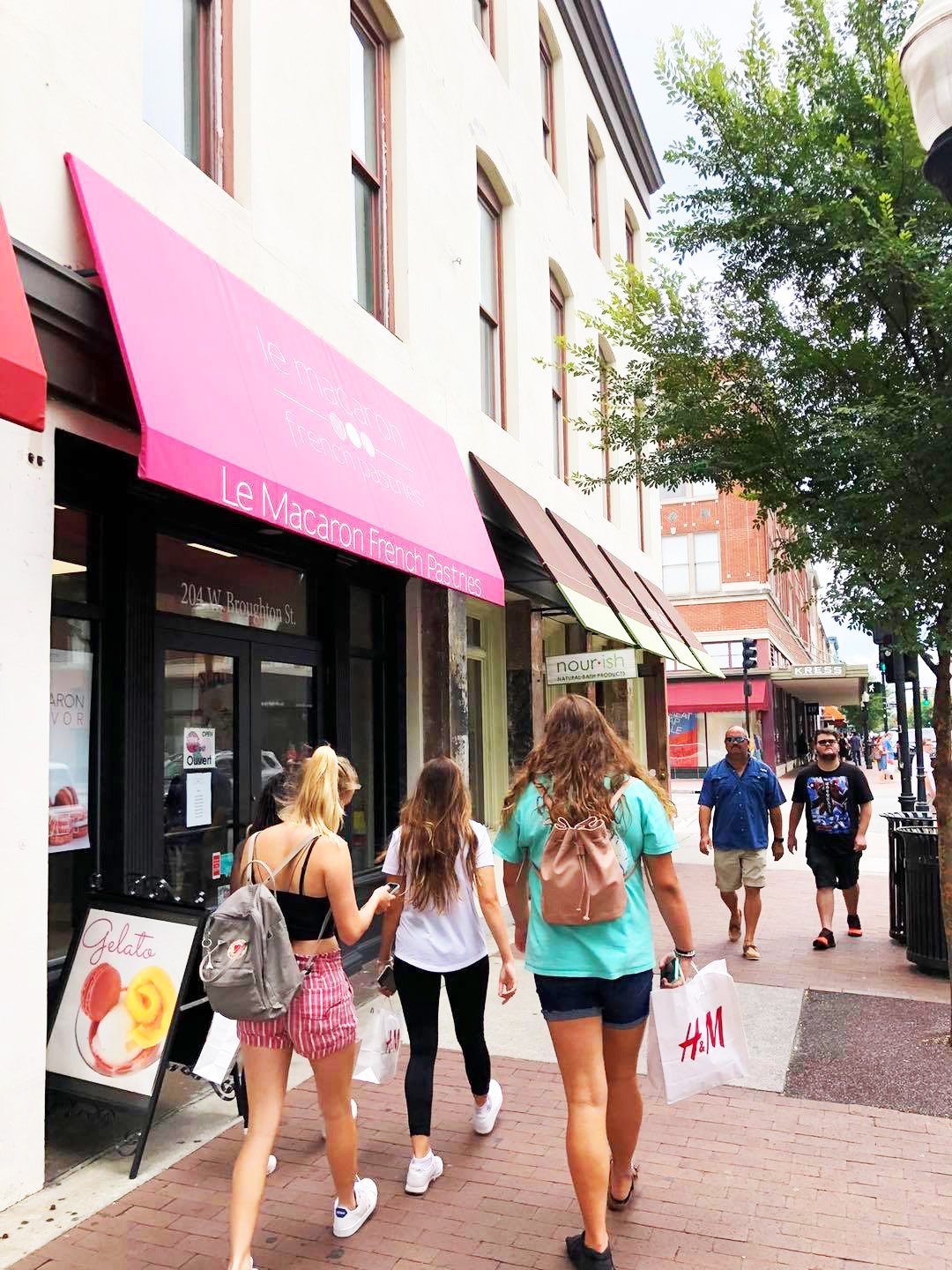 People walking on a city sidewalk past shops with awnings.
