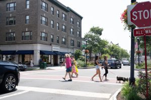 Pedestrians crossing a street in front of shops and apartment buildings.