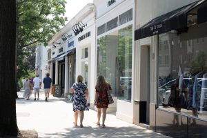 Two women walking past upscale shops on a sunny street.