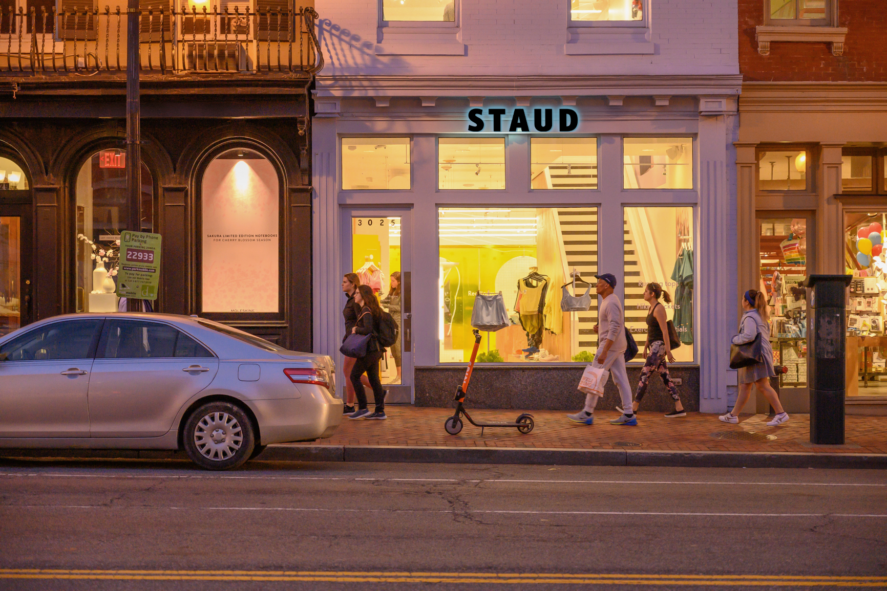 Staud storefront at dusk with pedestrians and a scooter.