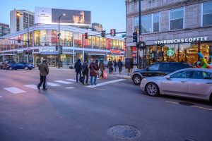 Pedestrians crossing a city street at dusk, with cars and shops in the background.