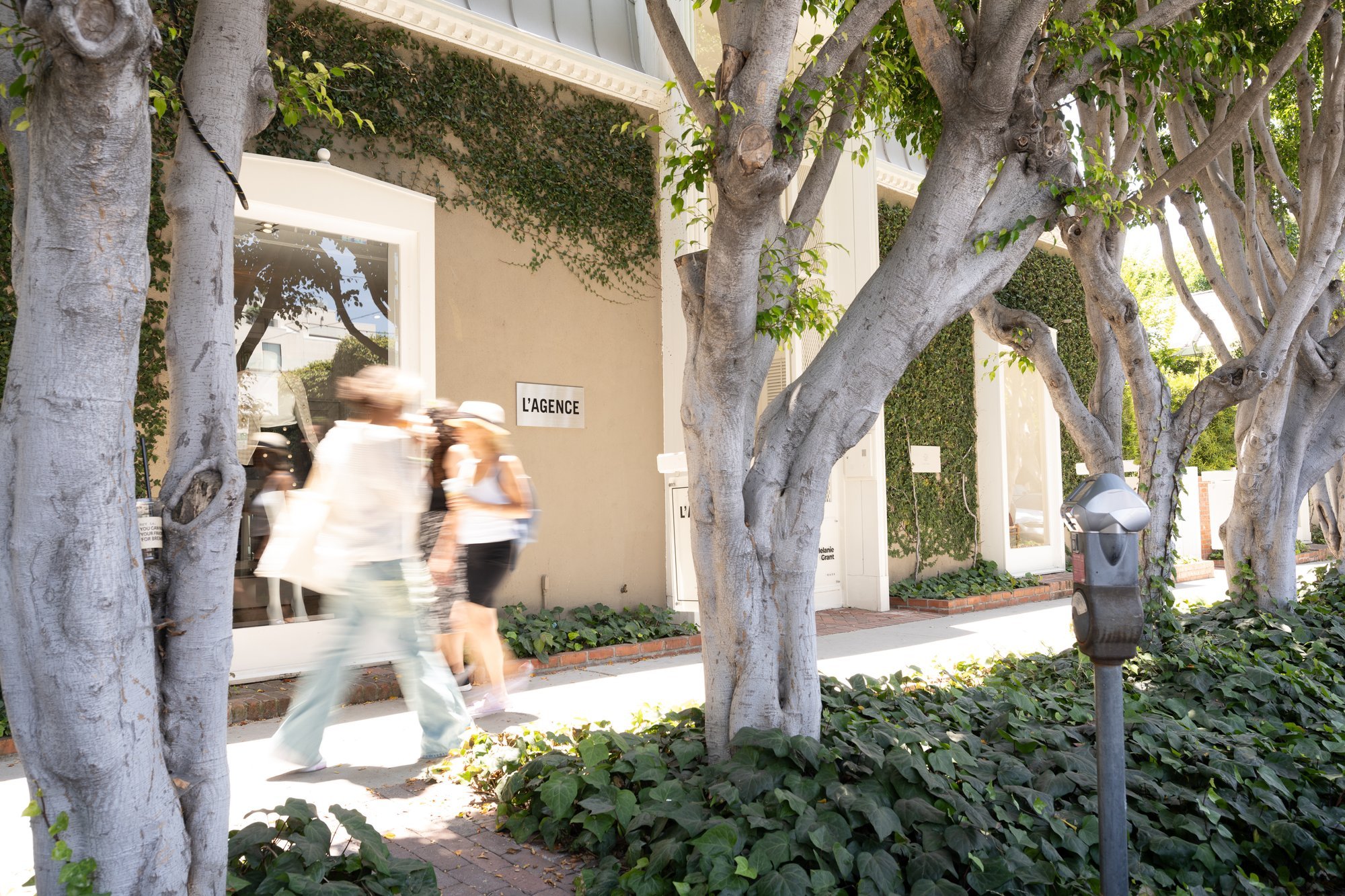 People walking past shops and trees on a sunny street.