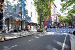 Pedestrians walking on a city street lined with shops and trees.