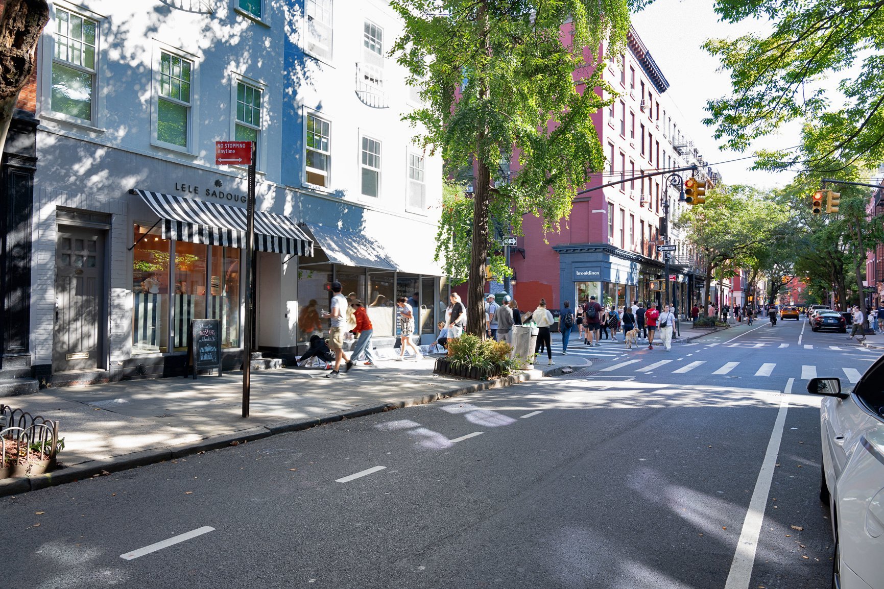 Pedestrians walking on a city street lined with shops and trees.