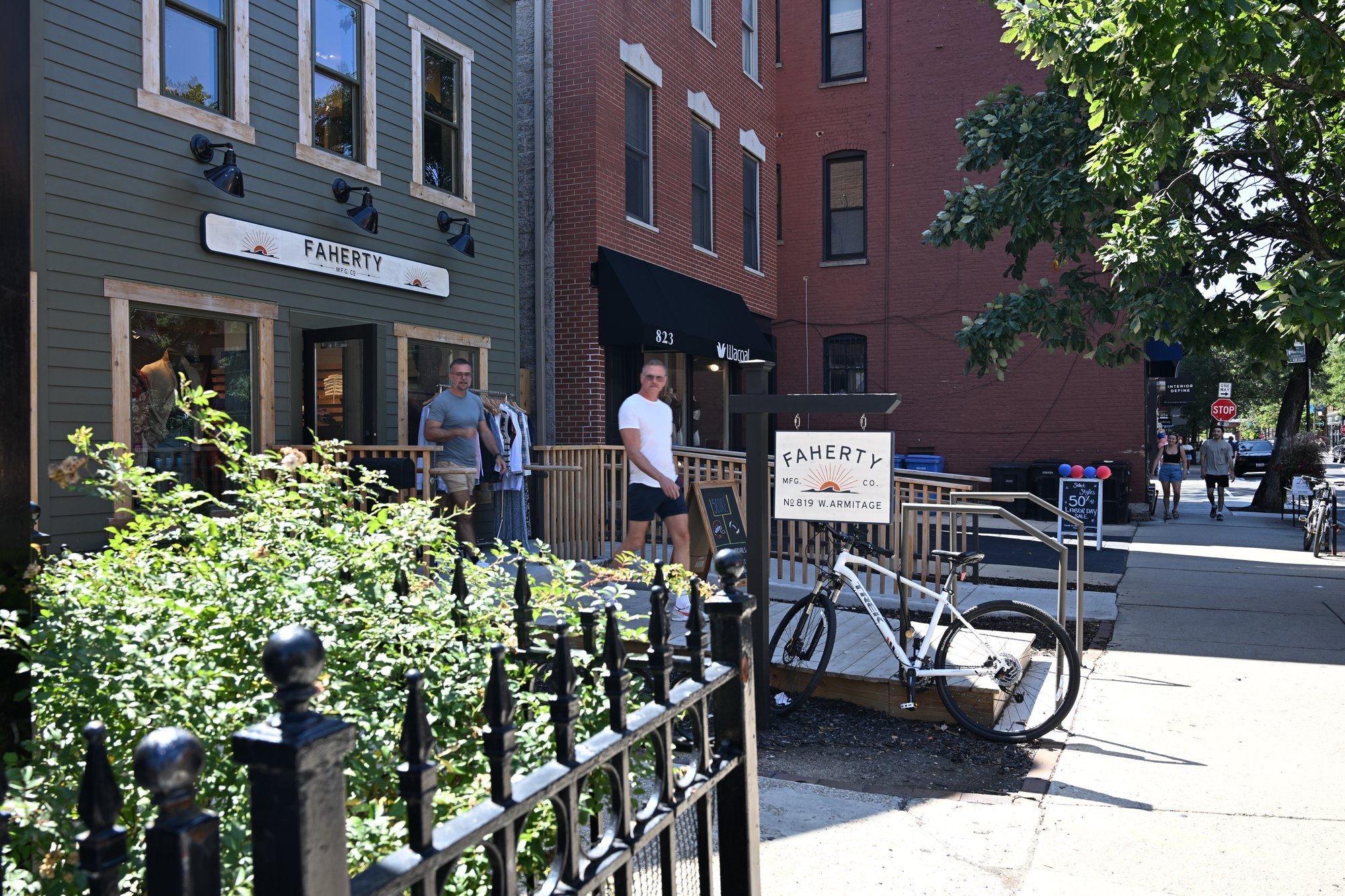 Two men standing outside a cafe, a bicycle, and city buildings in the background.