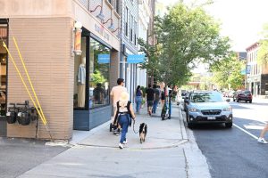 Person walking two dogs on a city sidewalk past shops.