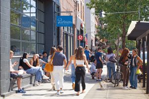 People walking and sitting on a city sidewalk outside shops.