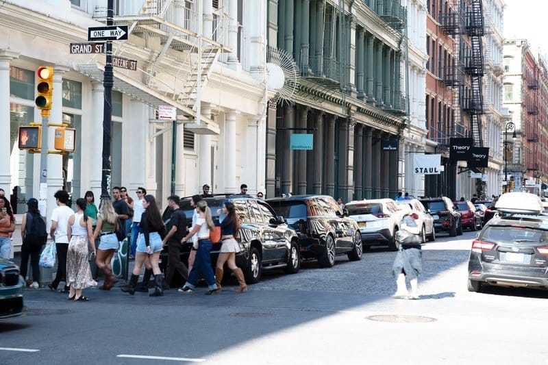 Pedestrians crossing a city street lined with parked cars and historic buildings.
