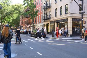Pedestrians and a cyclist crossing a city street lined with shops and trees.