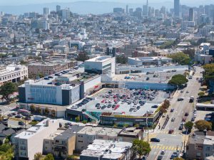 Aerial view of a shopping center and parking lot with San Francisco skyline in the background.