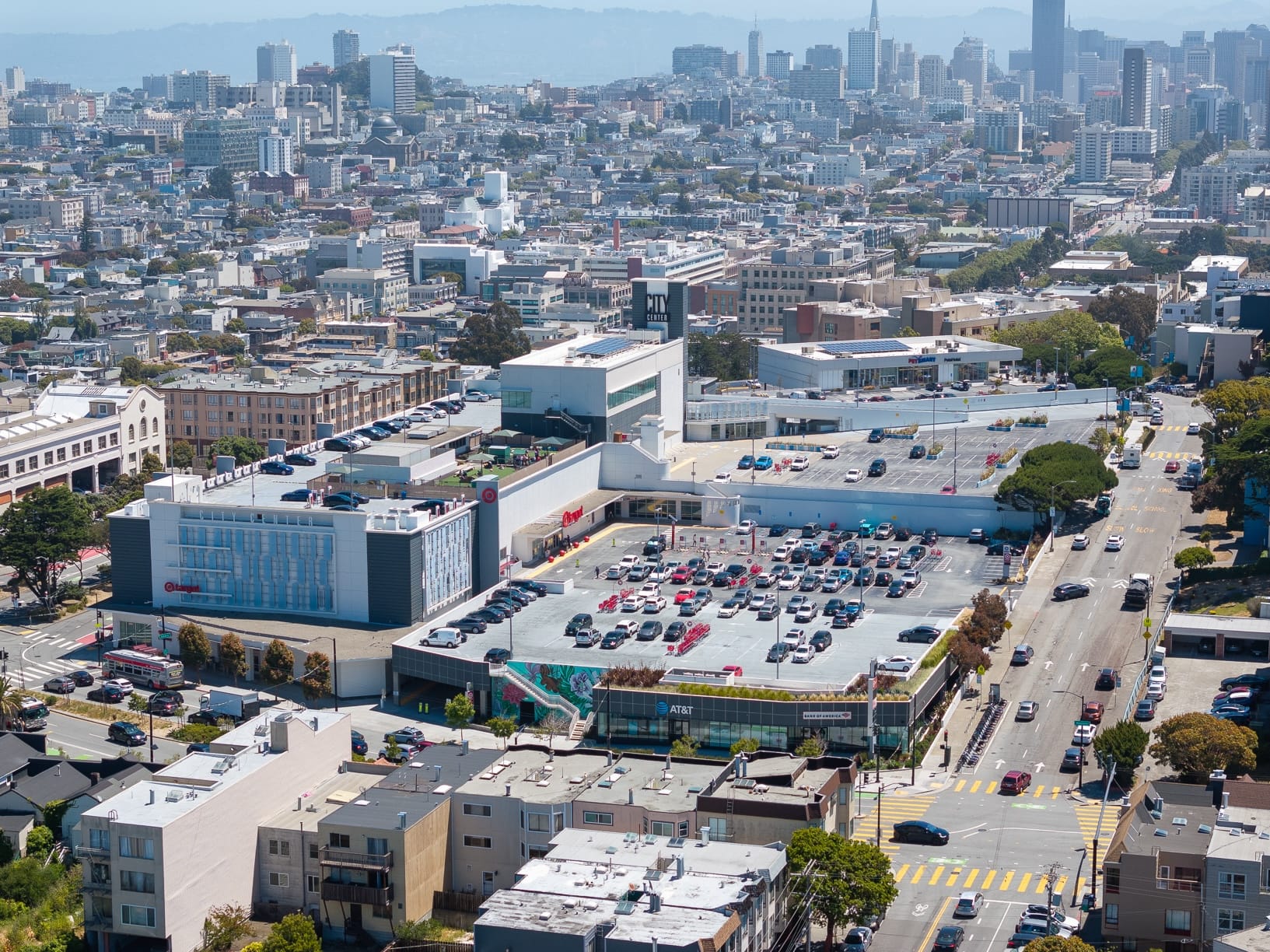 Aerial view of a shopping center and parking lot with San Francisco skyline in the background.