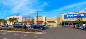 Sunny day at a shopping center with palm trees and parked cars.