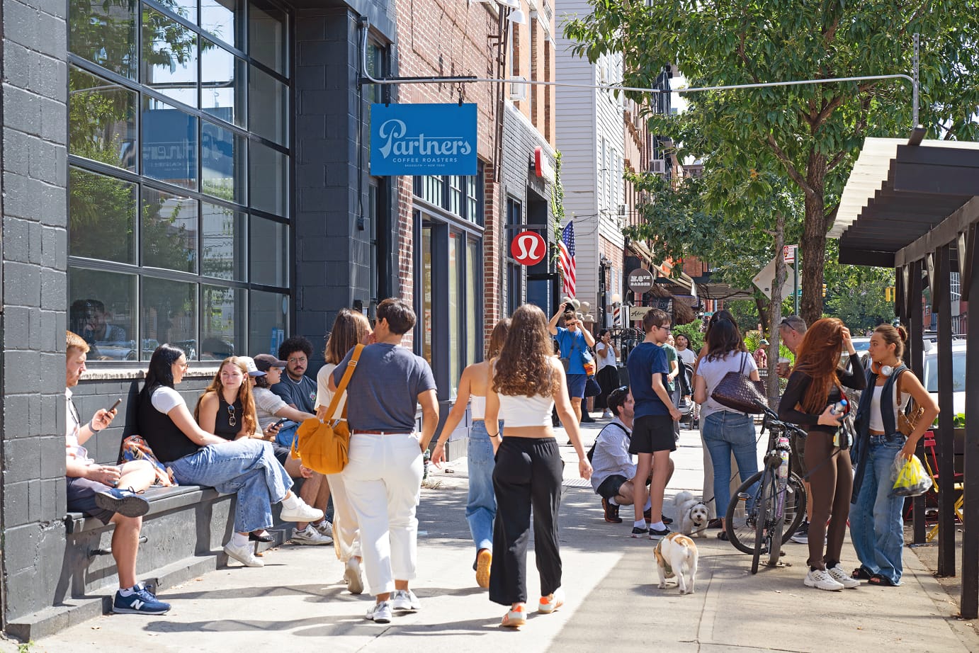 People walking and sitting outside shops on a sunny city street.